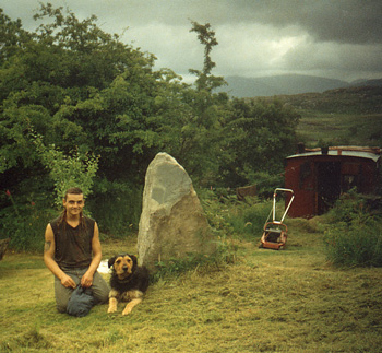 a young man and his dog by a standing stone in the hills, with their home close by.
